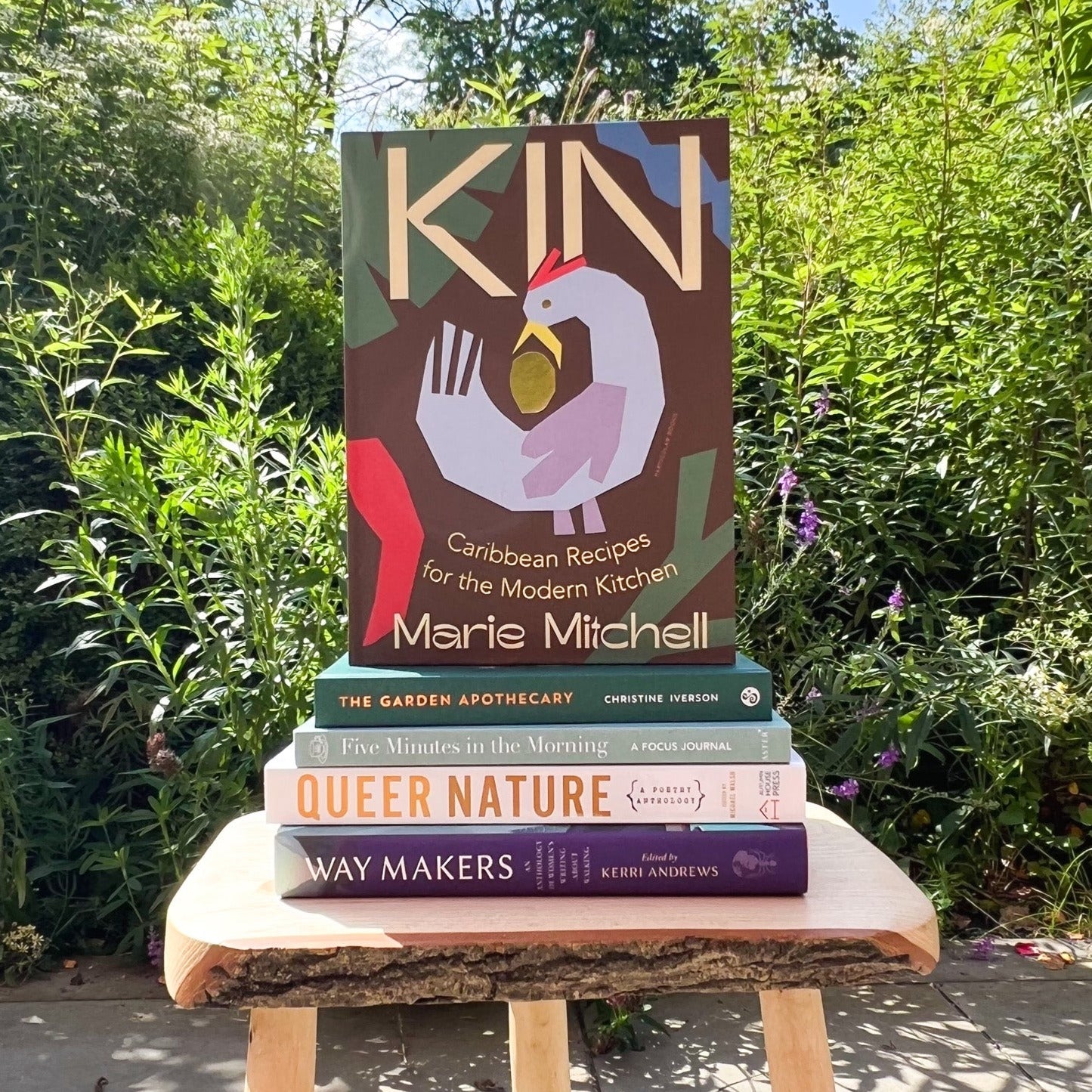 A carribbean cookbook stood upright on top of a stack of other books, in front of a garden setting.