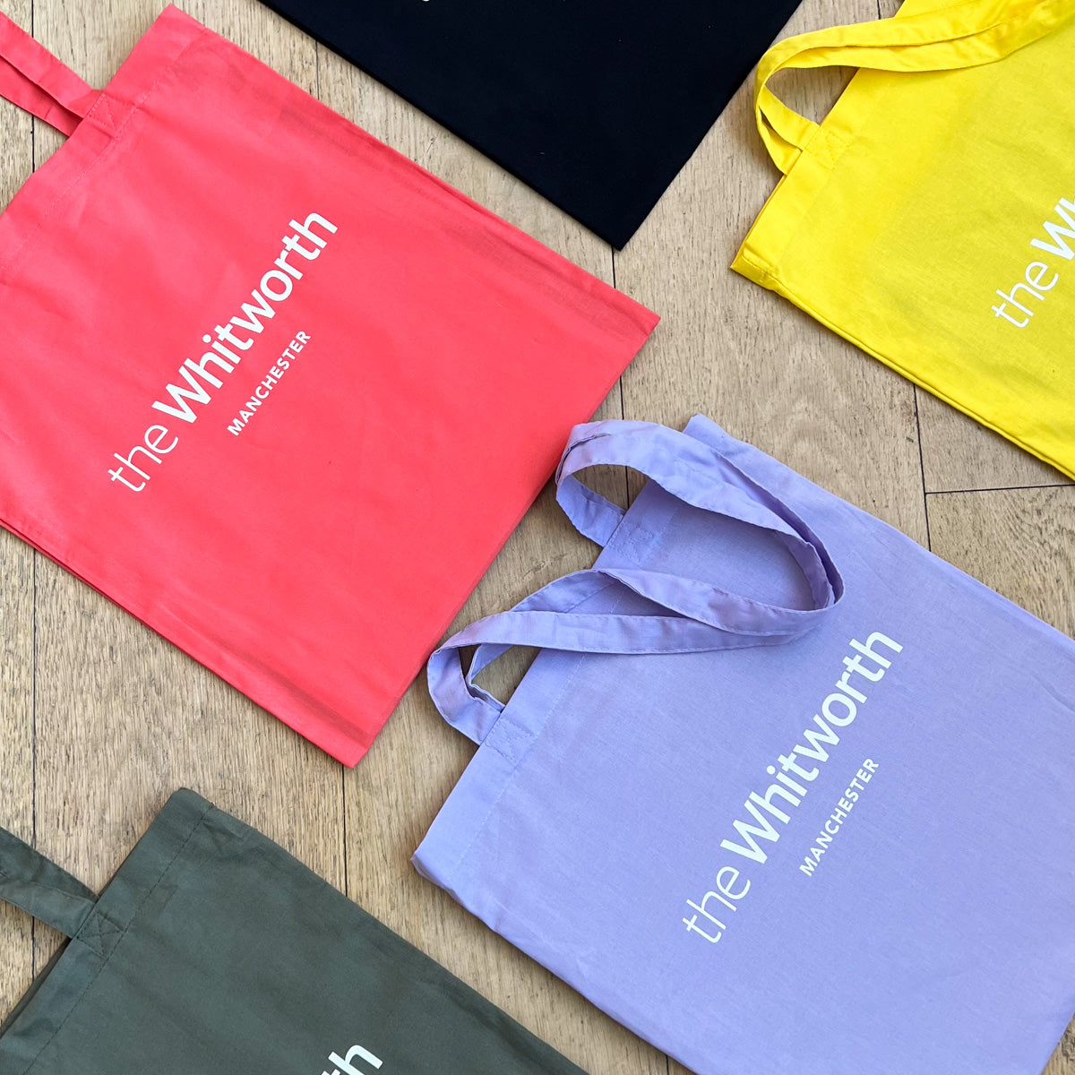 A series of tote bags laid in a grid format on a wooden surface. Each tote is a different colour and has the Whitworth logo at the centre.