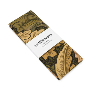 Tea towel featuring William Morris Acanthus leafy pattern. Wrapped around the tea towel is a belly band with the Whitworth's branding on it. White background.
