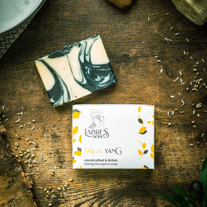 Birds-eye view of yellow packaging for Labre's Hope soap, with the bar of soap placed above. Both placed on a wooden surface.