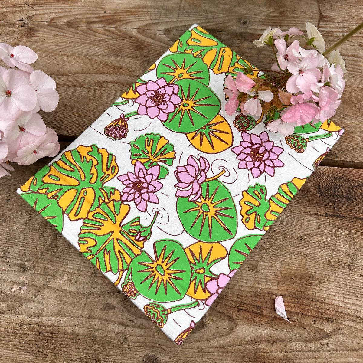 Notebook seen at an angle on a wooden surface. Water lily illustrations in pink, green and yellow are printed across the notebook.