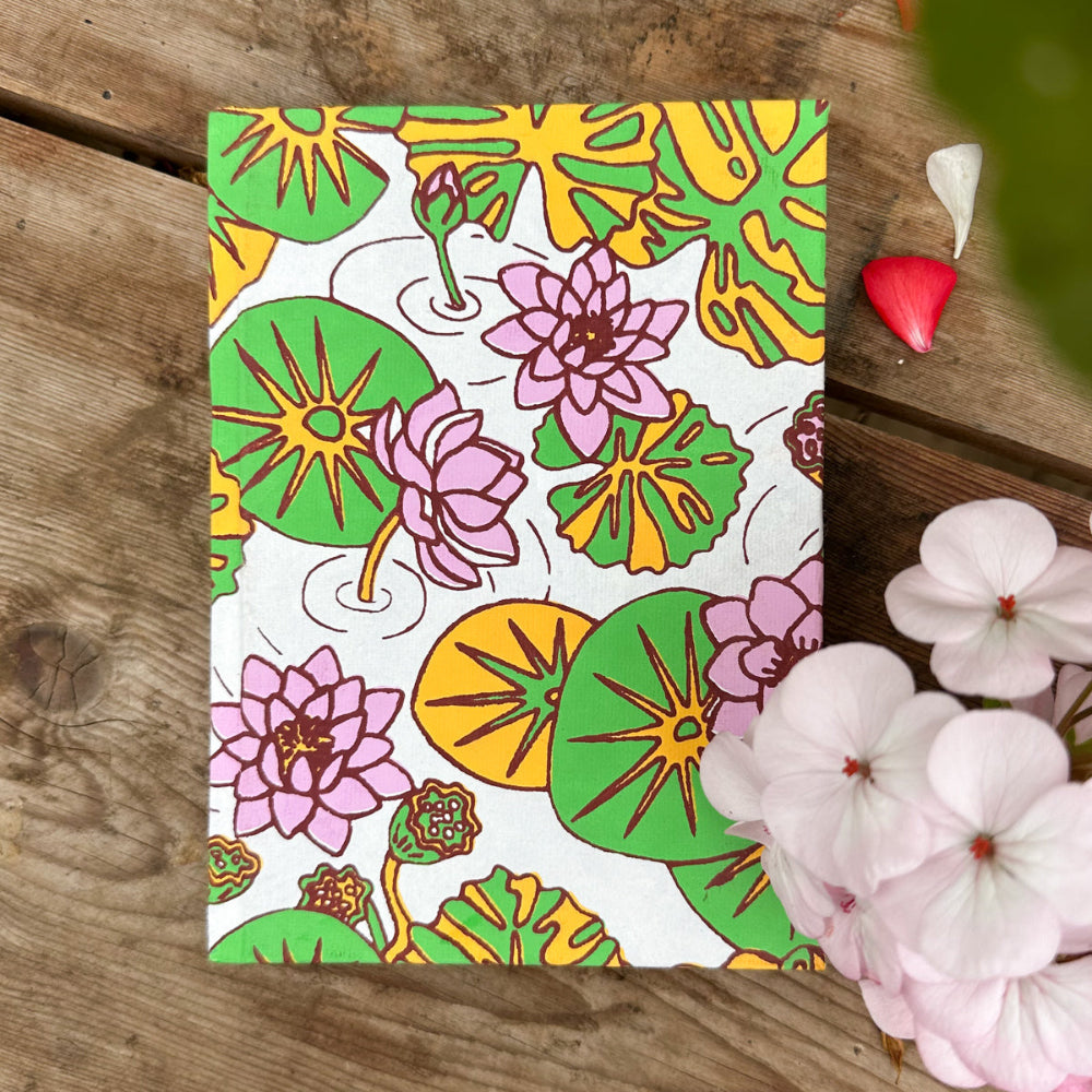 Notebook on wooden surface with pelagonium flowers to the right. The notebook is printed with water lily illustrations.