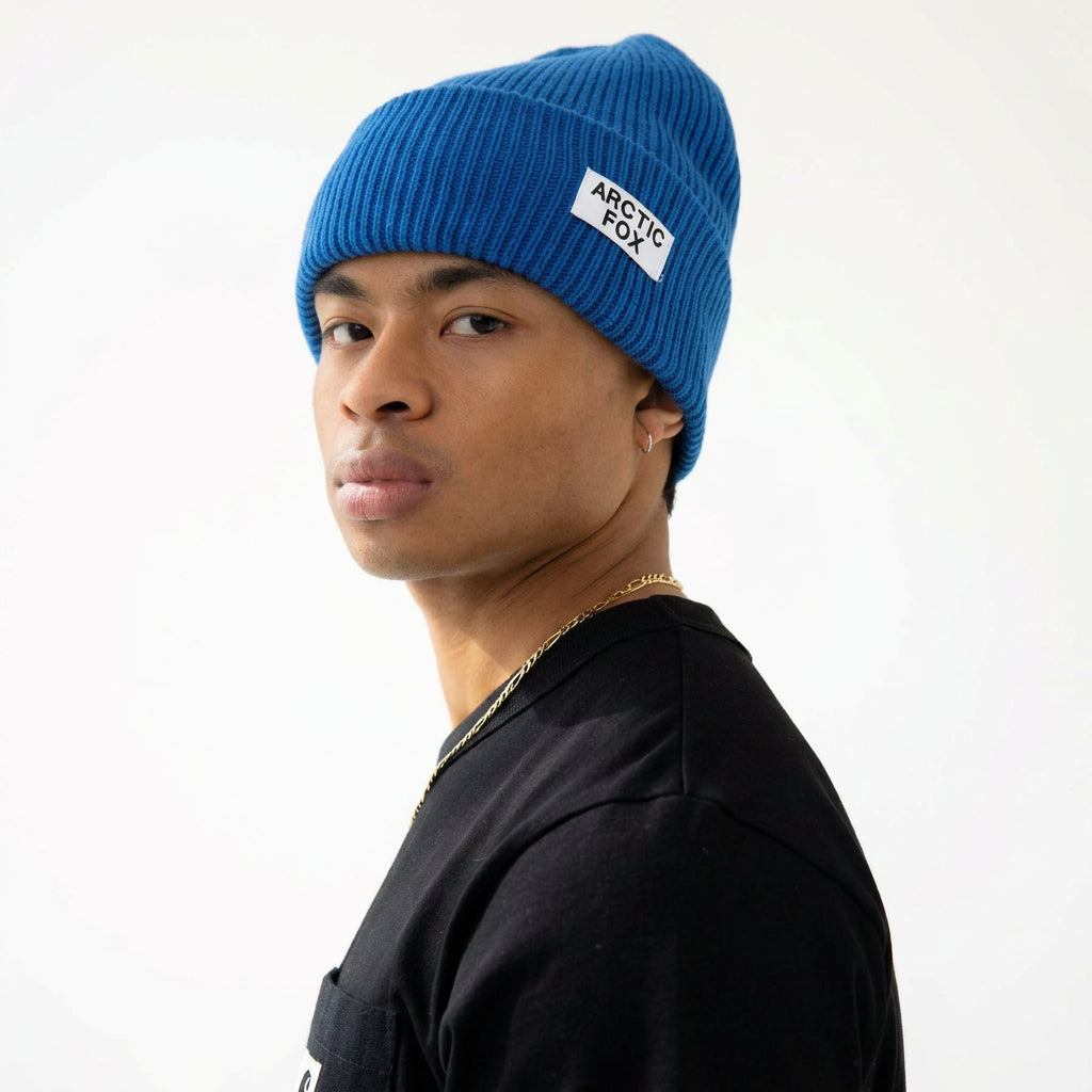A man wearing a bright blue ribbed beanie with a white 'Arctic Fox' logo patch and a black t-shirt, shown in profile against a plain white background.