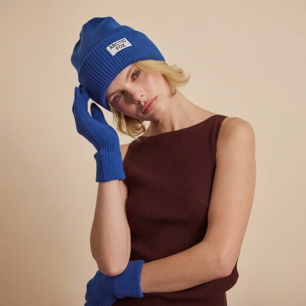A model wearing a royal blue ribbed knit beanie with a white 'Arctic Fox' logo patch, matching blue knit gloves, and a dark brown ribbed knit top, posed against a neutral light brown background.