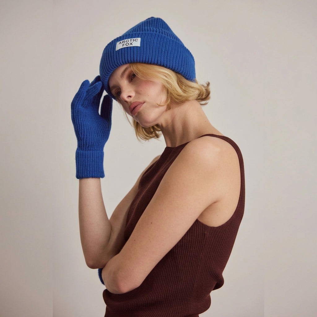 A model wearing a vibrant blue knit beanie with a white 'Arctic FOX' label and matching blue knit gloves, paired with a dark brown ribbed tank top, set against a plain light brown studio background.