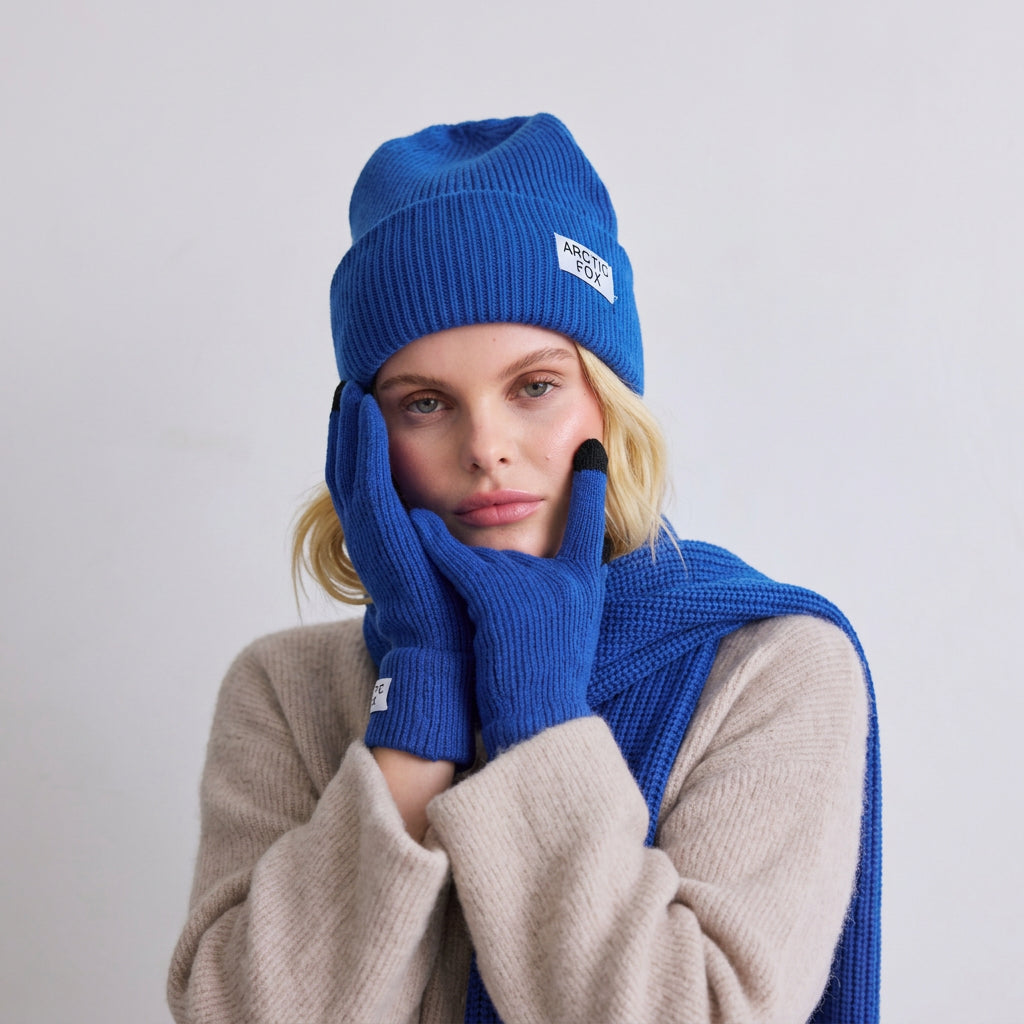 A woman wearing a bright blue ribbed knit beanie, matching scarf, and gloves with black touchscreen-compatible fingertips, over a beige sweater, photographed against a light gray studio background.