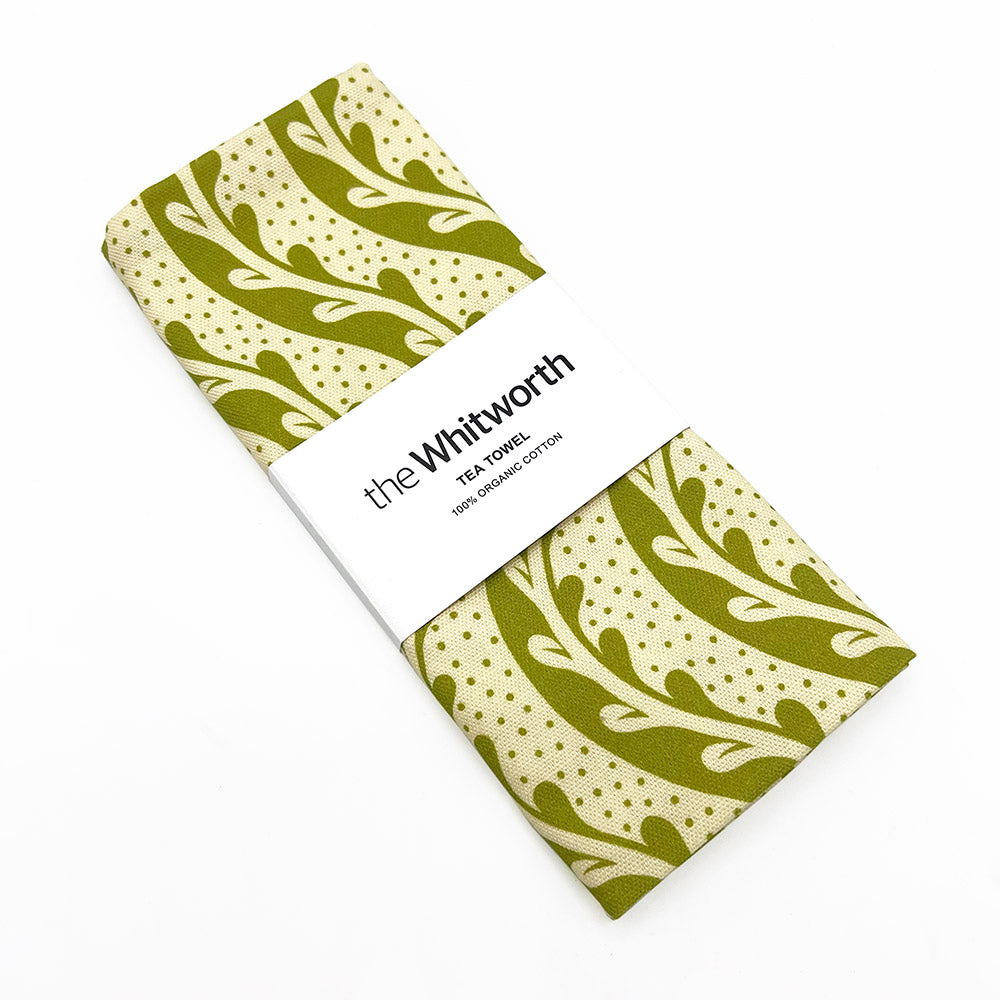 Green patterned tea towel folded up with a Whitworth branded white belly band. Presented on a white background.