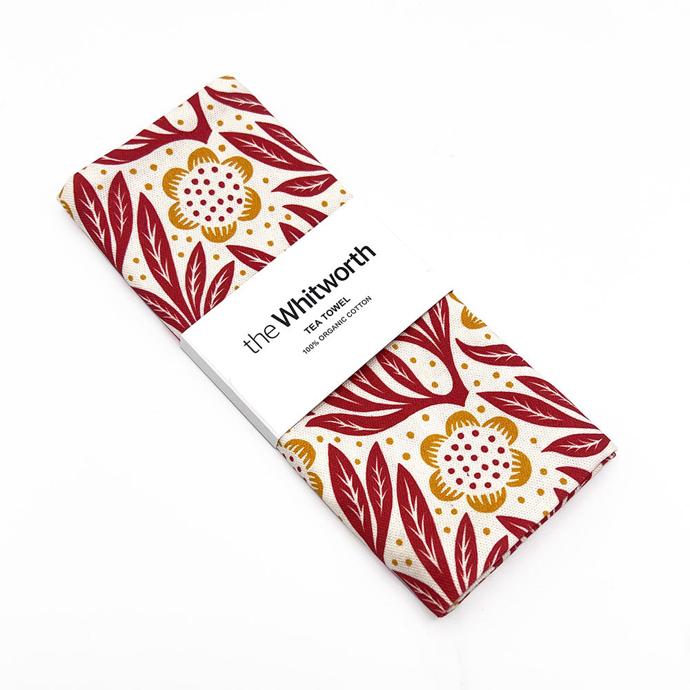Red patterned tea towel folded up with a Whitworth branded white belly band. Presented on a white background.
