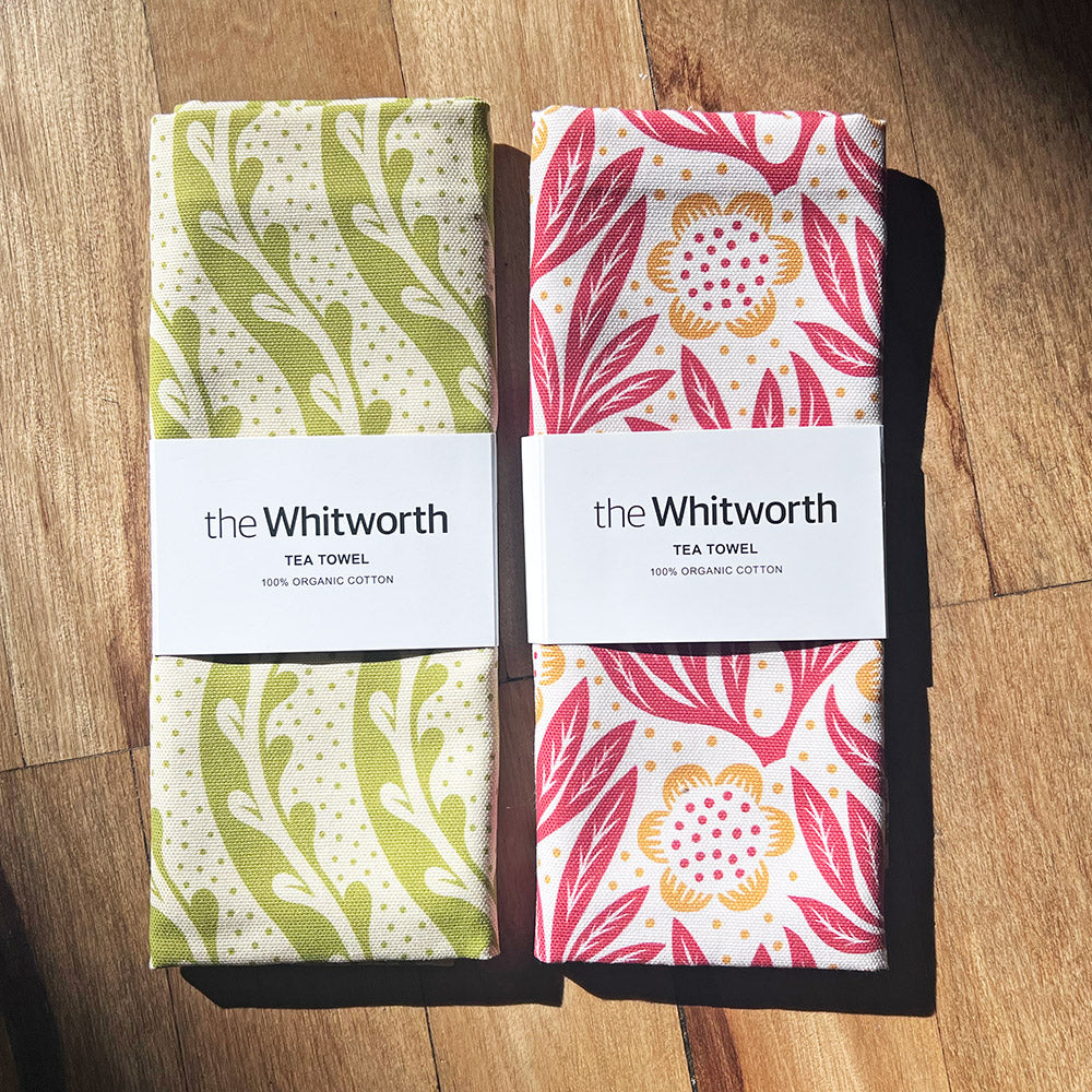 Two patterned tea towel folded up with a Whitworth branded white belly band - sat site by side on a wooden surface.