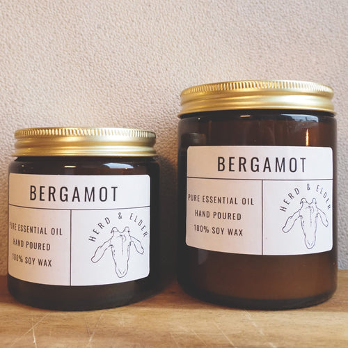 Both sizes of the bergamot candle standing next to each other with the smaller to the left. The candles are of brown glass with gold metal lids. Simple paper branding stickers on the front in white and black text.