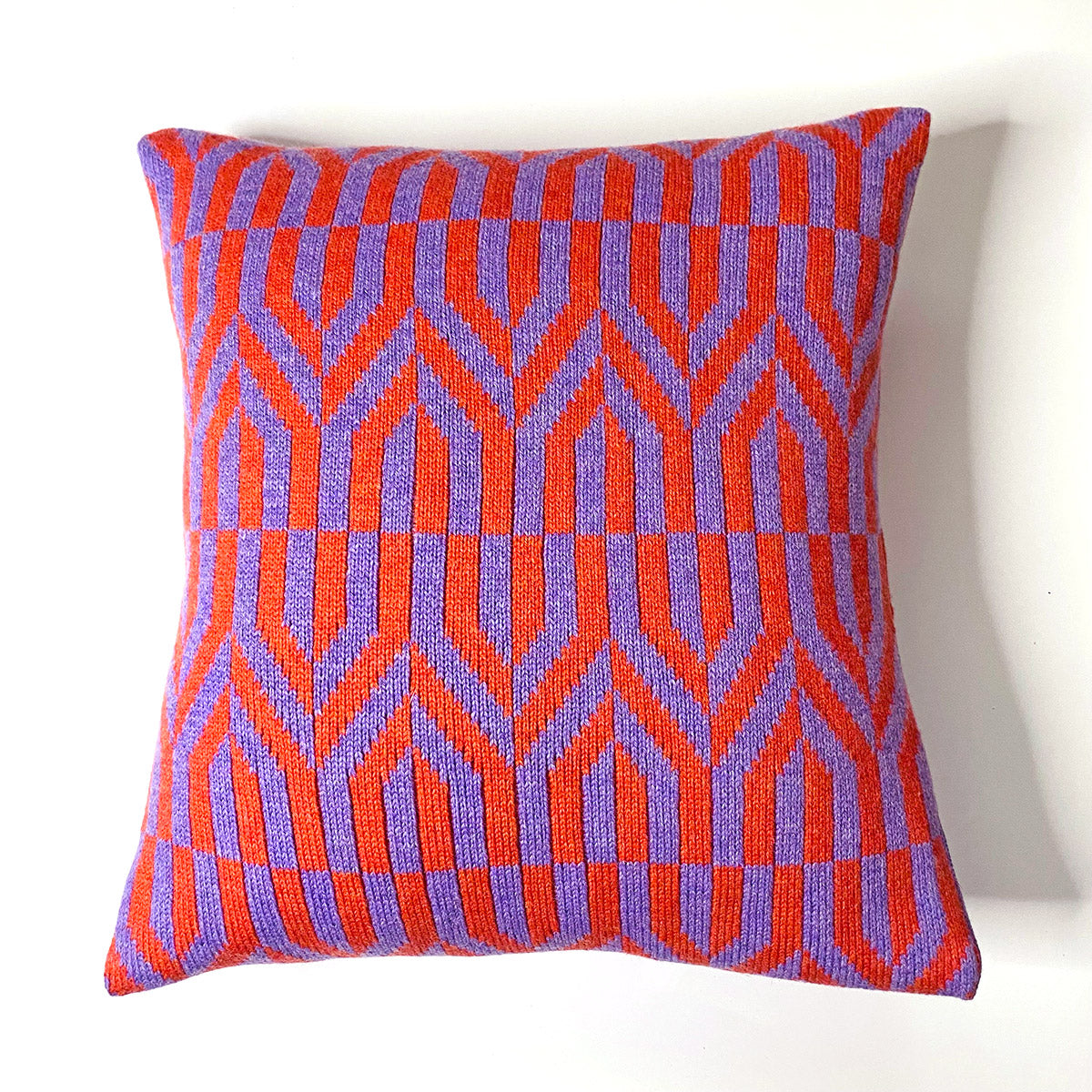 A square pillow with a bold orange and purple geometric pattern. The pattern features interlocking shapes that resemble arrows pointing in different directions.