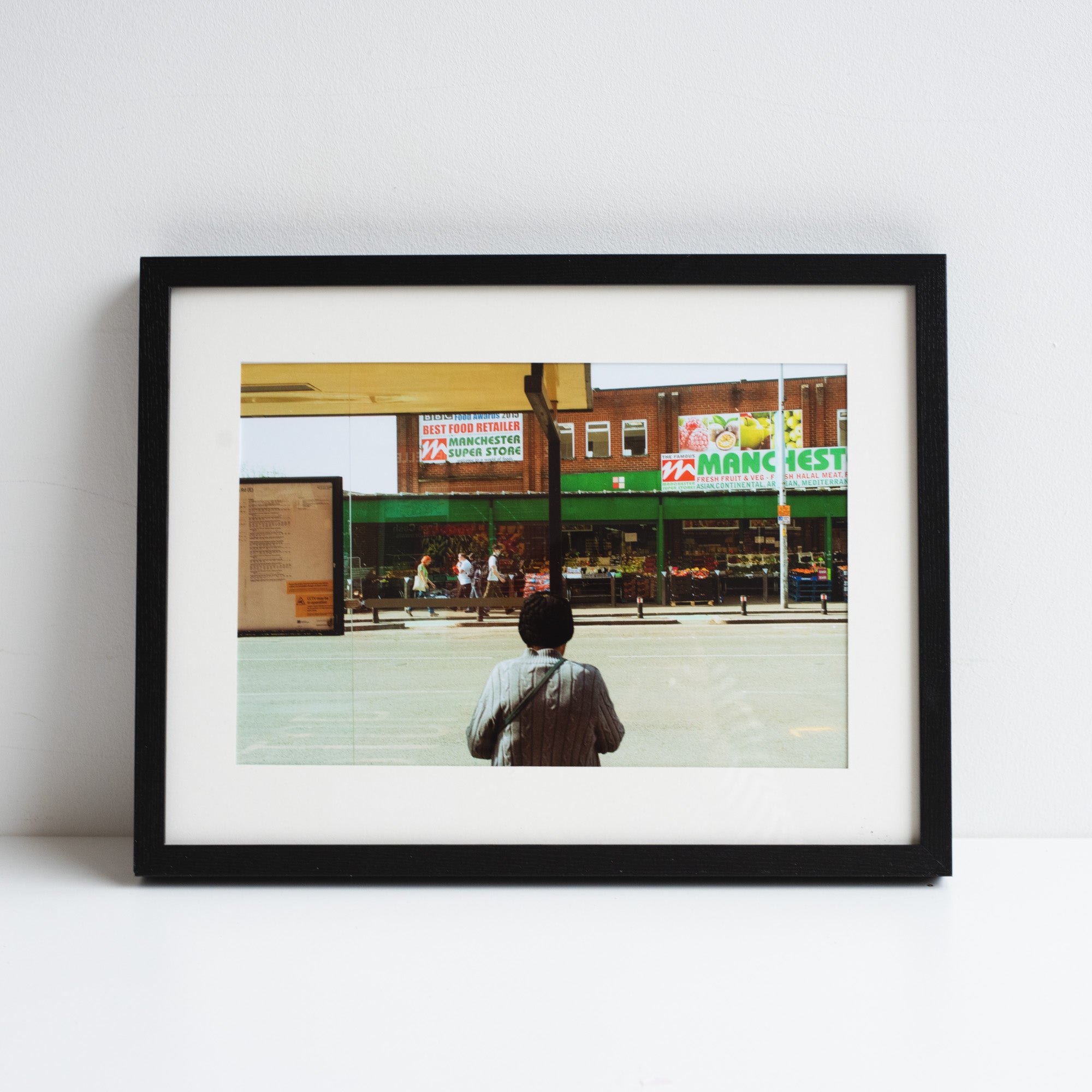 A printed reproduction of a photograph by Chris Naylor of a man looking our to a groceries shop in Manchester. Placed in a black frame leaning against a white wall.