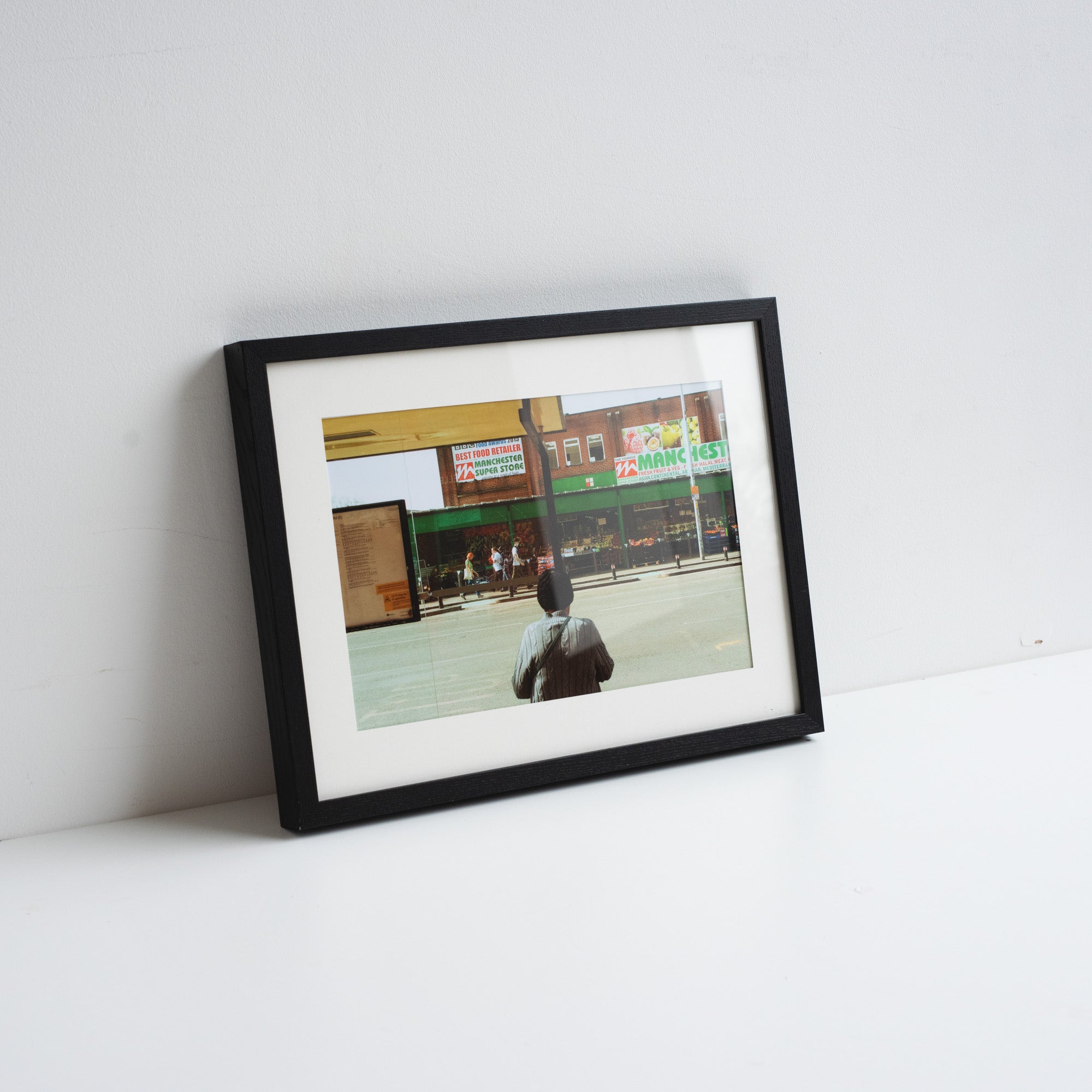 A printed reproduction of a photograph by Chris Naylor of a man looking our to a groceries shop in Manchester. Placed in a black frame leaning against a white wall.