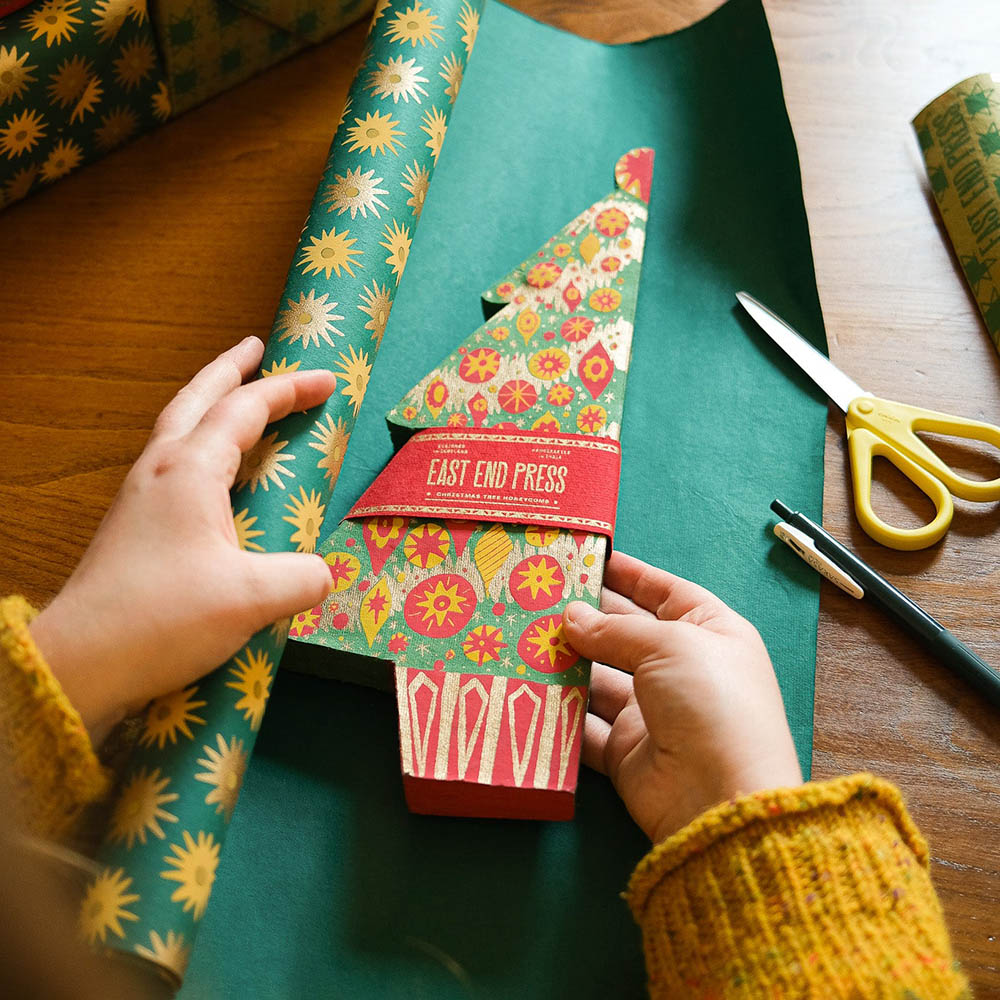 Person holding a floral-patterned gift box with 'East End Press' on a wooden table with wrapping paper and scissors.