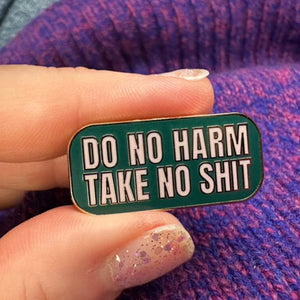 Person holding an enamel pink with pink 'DO NO HARM TAKE NO SHIT' on a forest green background.