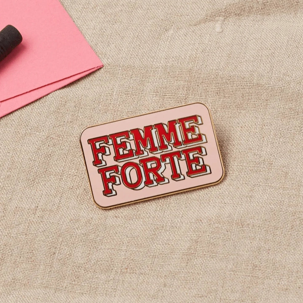 A "Femme Forte" pin rests on a neutral surface next to a pink card and black item.