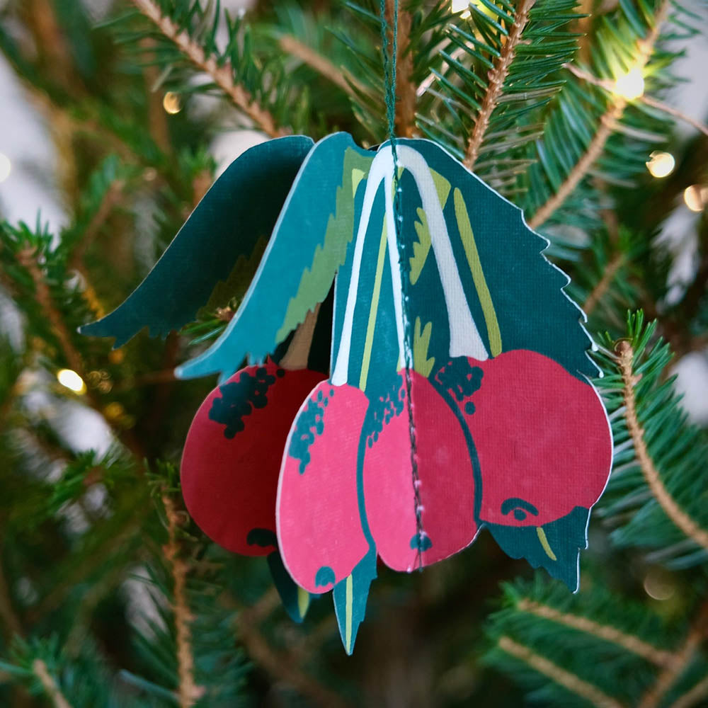 Decorative ornament with pink berries and green leaves hanging on a Christmas tree.