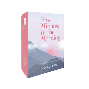 The image shows a box of cards called "Five Minutes in the Morning: A Focus Deck." The box is a light blue colour with a pink sunset and mountains in the background. The text on the box is white and says "Five Minutes in the Morning" and "50 Cards to Change Your Day."