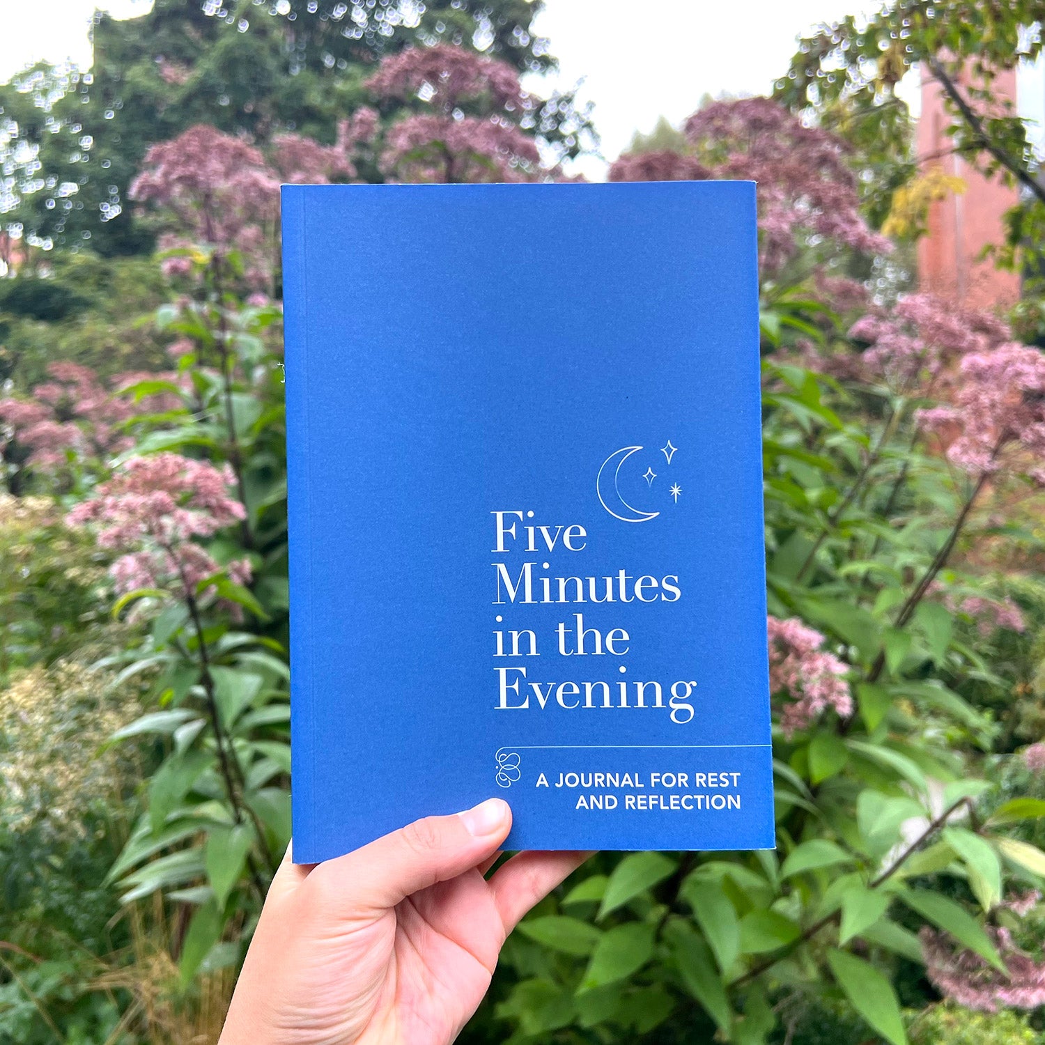 A person's hand holding a blue journal with the title "Five Minutes in the Evening: A Journal for Rest and Reflection." The background is blurred and shows a green garden with flowers.