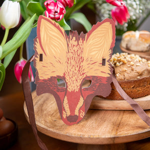 Fox mask on a copping board in front of cake and flowers.