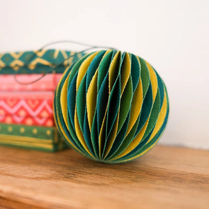 Paper bauble ornament made from multiple layers of paper in shades of green and yellow. The bauble is resting on a wooden surface, with a stack of gift boxes visible in the background. 