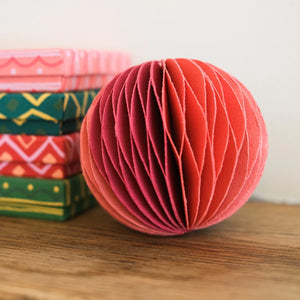 Paper bauble ornament made from multiple layers of paper in shades of red and pink. The bauble is resting on a wooden surface, with a stack of gift boxes visible in the background. 