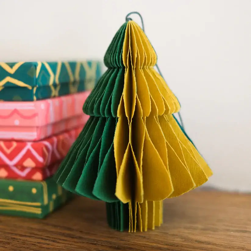 A close-up photo of a green and yellow paper Christmas tree ornament. The ornament is folded into a honeycomb shape and stands upright on a wooden surface. There are other gift boxes in the background.