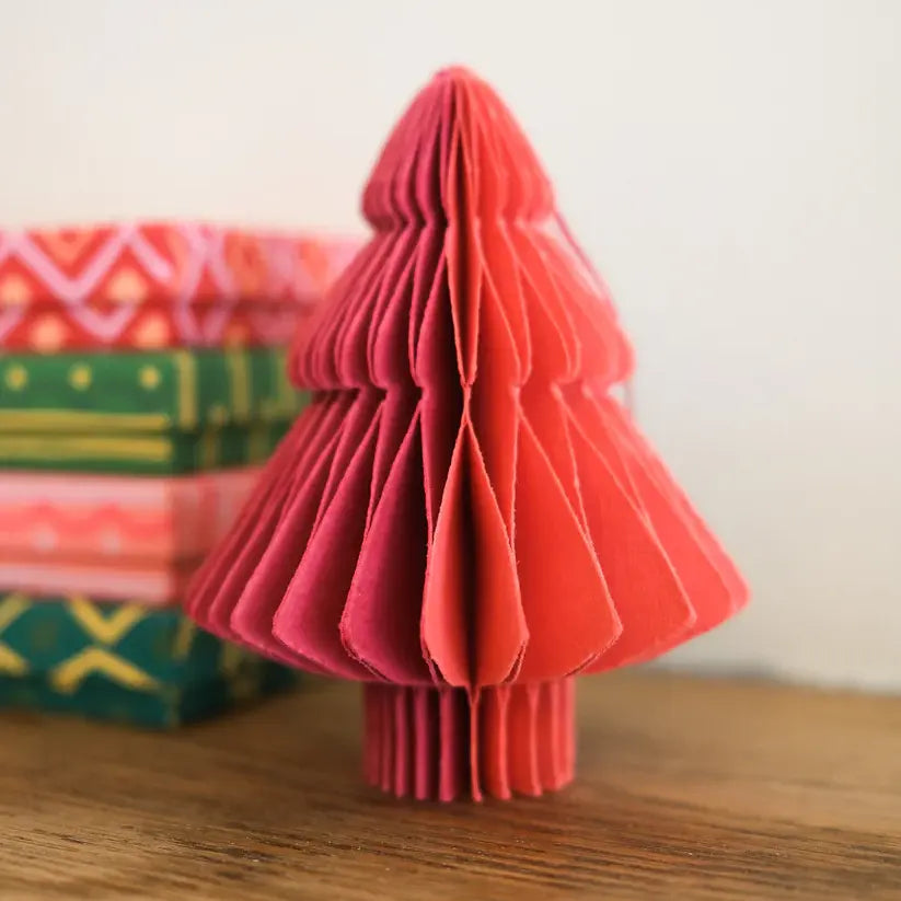 A close-up photo of a red and pink paper Christmas tree ornament. The ornament is folded into a honeycomb shape and stands upright on a wooden surface. There are other gift boxes in the background.