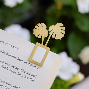 The image shows a gold bookmark shaped like a monstera plant, marking a page in a book.