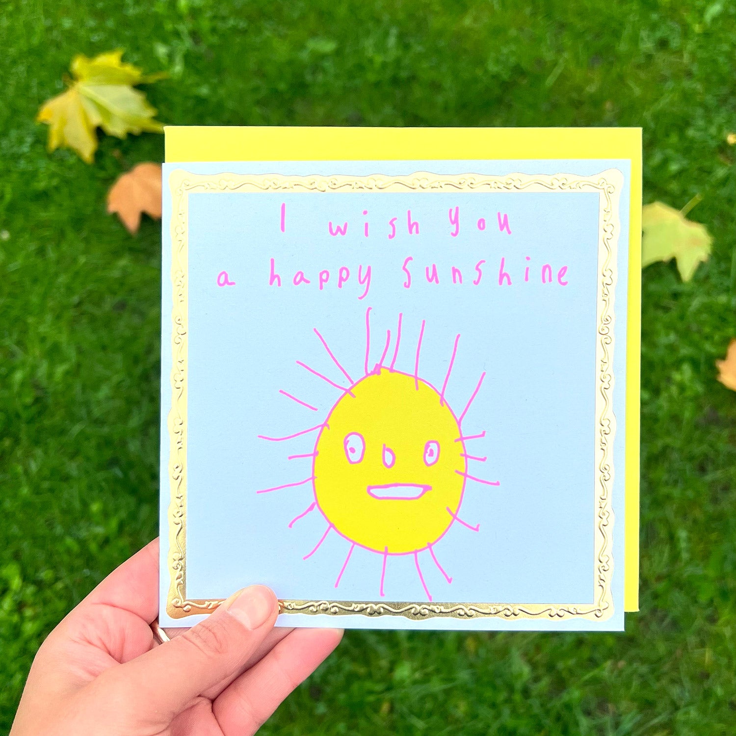 A person holding a greetings card against a grass/leaves backdrop. It is a white greeting card with a gold border. In the centre is a child-like drawing of a yellow sun with a smiling face and pink rays. Text in pink reads "I wish you a happy sunshine."