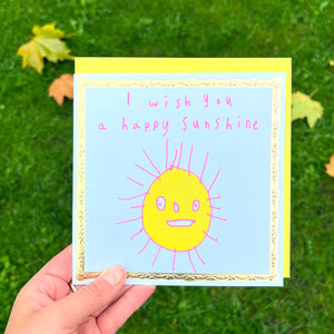 A person holding a greetings card against a grass/leaves backdrop. It is a white greeting card with a gold border. In the centre is a child-like drawing of a yellow sun with a smiling face and pink rays. Text in pink reads "I wish you a happy sunshine."