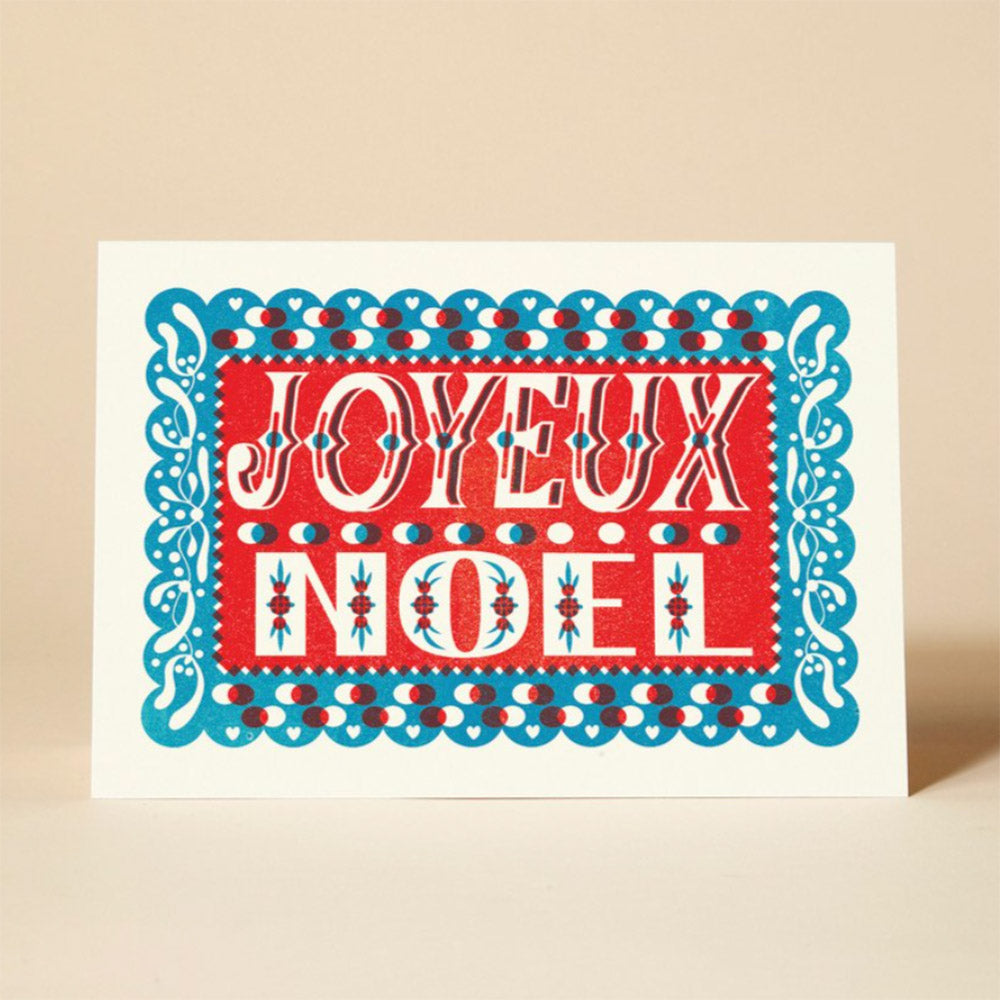 A vibrant Christmas card featuring the French phrase "JOYEUX NOEL" in red and blue lettering, surrounded by intricate paper cut-out designs. 