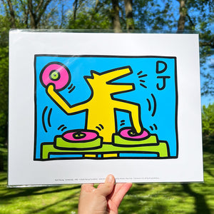A person holding arinted reproduction of an artwork by Keith Haring titled DJ, in front of some trees. The dog in is situated behind a turn table and holds a record in one hand.