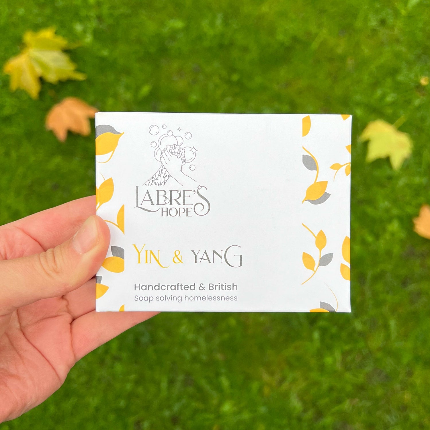 A person holding the packaging for a bar of soap above some autumn leaves