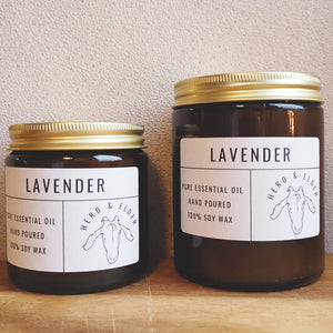 Both sizes of the lavender candle standing next to each other with the smaller to the left. The candles are of brown glass with gold metal lids. Simple paper branding stickers on the front in white and black text.