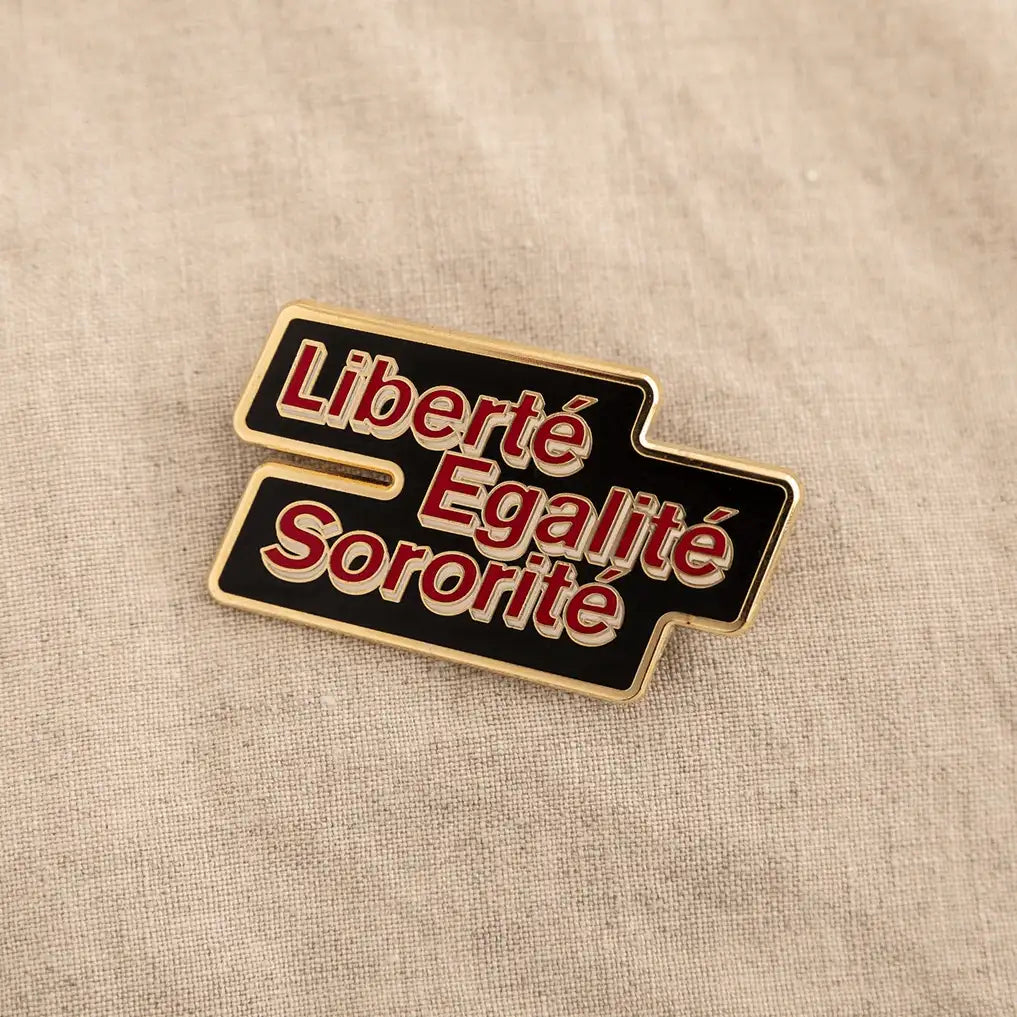 A black enamel pin with the words "Liberté," "Egalité," and "Sororité" written in gold. The words are arranged in a rectangular shape with rounded corners.