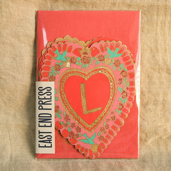 Front panel of a concertina greetings card in the shape of a love heart with the letter L in the middle. Placed on top of a red envelope.