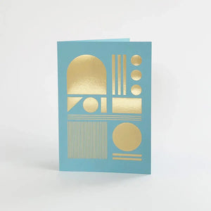 A light blue greeting card with gold foil geometric shapes, including circles, rectangles, and arches.