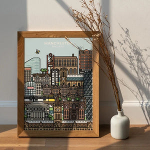 A framed print of a colorful, illustrated Manchester skyline. The print features iconic buildings in a detailed, almost pixelated style. The text "Manchester, England" is displayed at the top of the image. The print is shown resting on a wooden surface with dried flowers in a vase nearby.