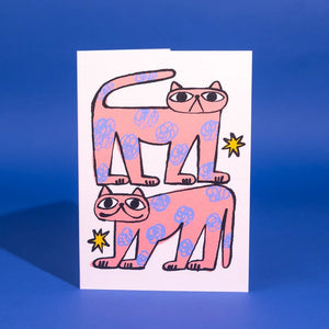 This is a white greetings card with a drawing of two pink cats with blue flower-like spots and yellow stars around them.