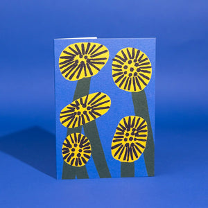 A blue greetings card with a pattern of five stylised yellow flowers with dark centres and dark green stems.