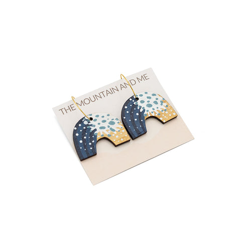 Arch-shaped wooden earrings wive wave painted detail, photographed against branded backing card on a white backdrop.