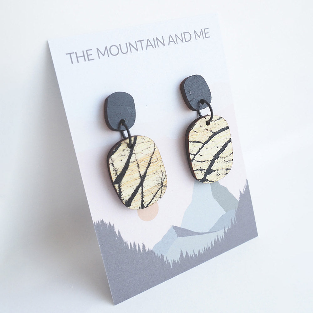 Pair of dangly wooden earrings with brand-like pattern on a 'The Mountain and Me' branded card with a mountain design.