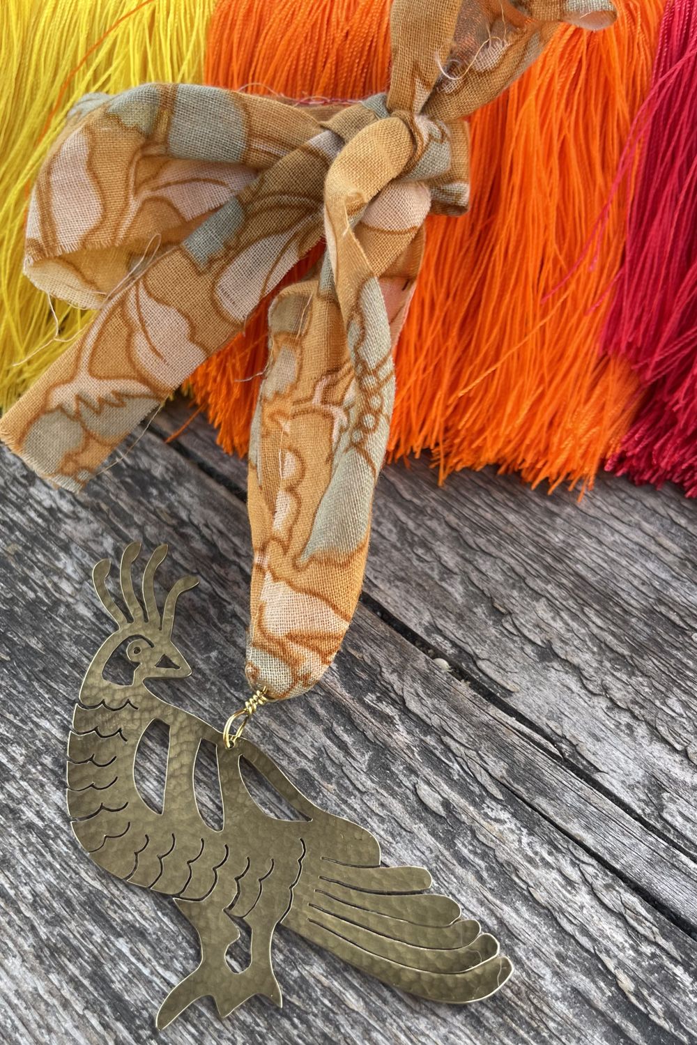 Brass peacock decoraitons with a fabric ribbon and seen on a wooden surface with colorful yarn in the background.