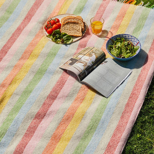 Colourful striped picnic blanket with food and a magazine on a grassy surface