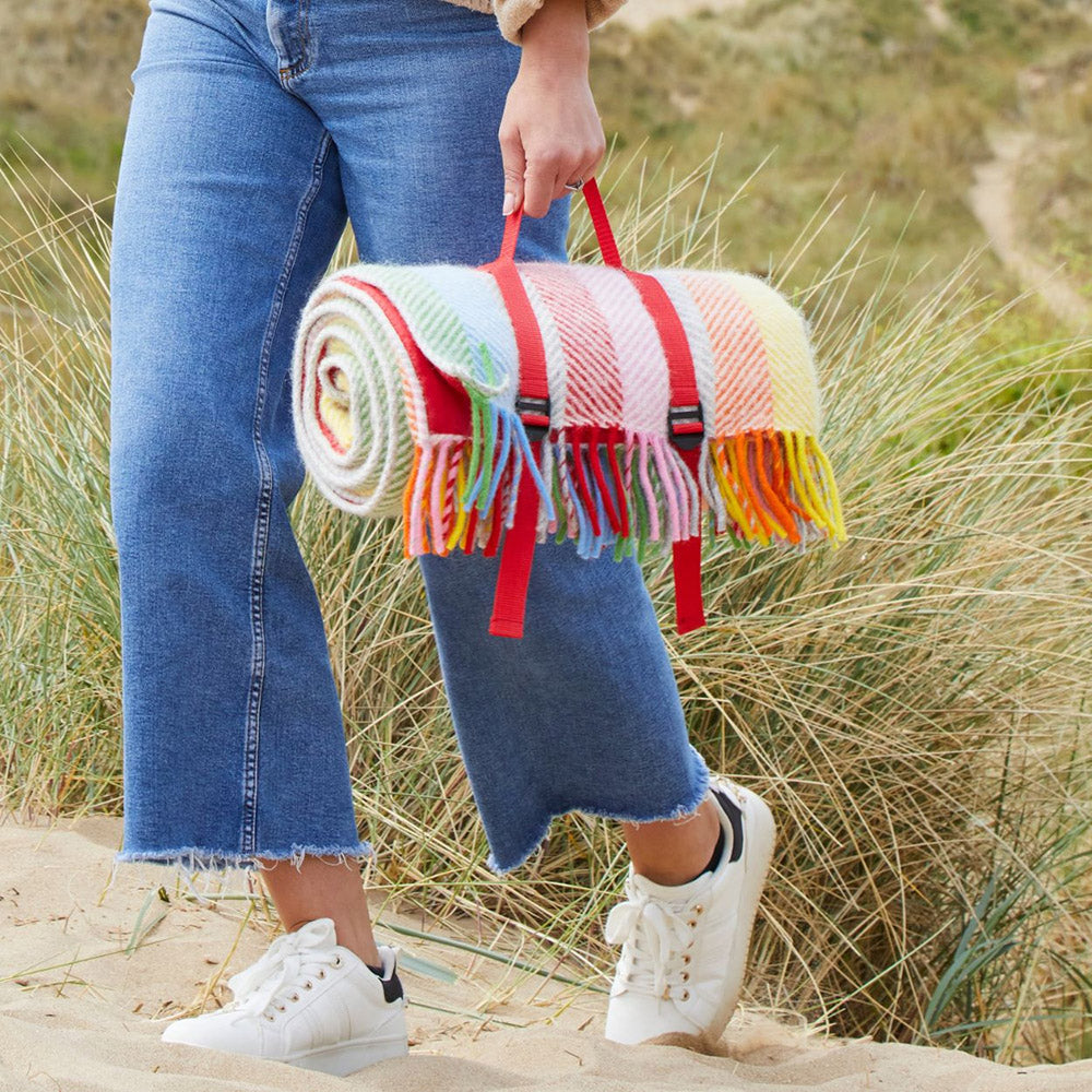 Someone holding a rainbow striped picnic rug with a red strap in front of grass
