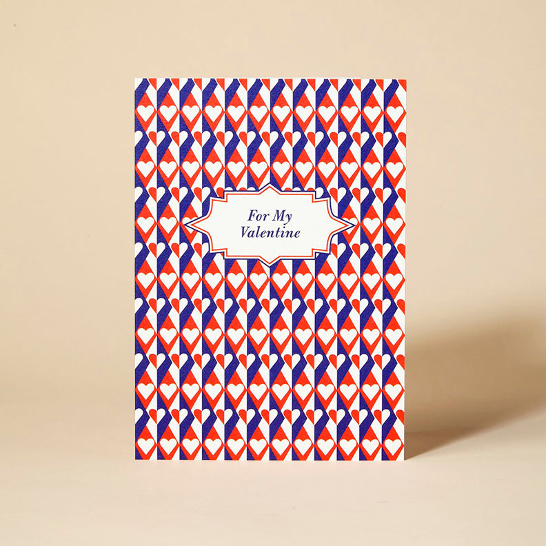 Card with the words For My Valentine at the centre. The card has a blue, red and white heart repeat pattern.