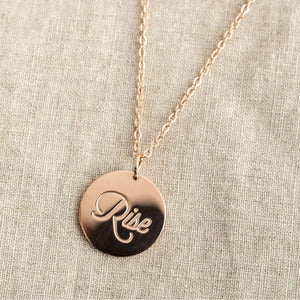 A rose gold necklace with a circular pendant engraved with the word "Rise."