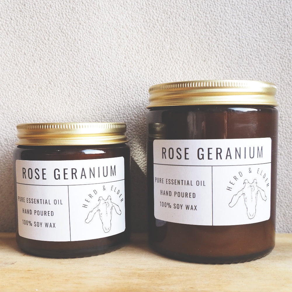 Both sizes of the rest candle standing next to each other with the smaller to the left. The candles are of brown glass with gold metal lids. Simple paper branding stickers on the front in white and black text.