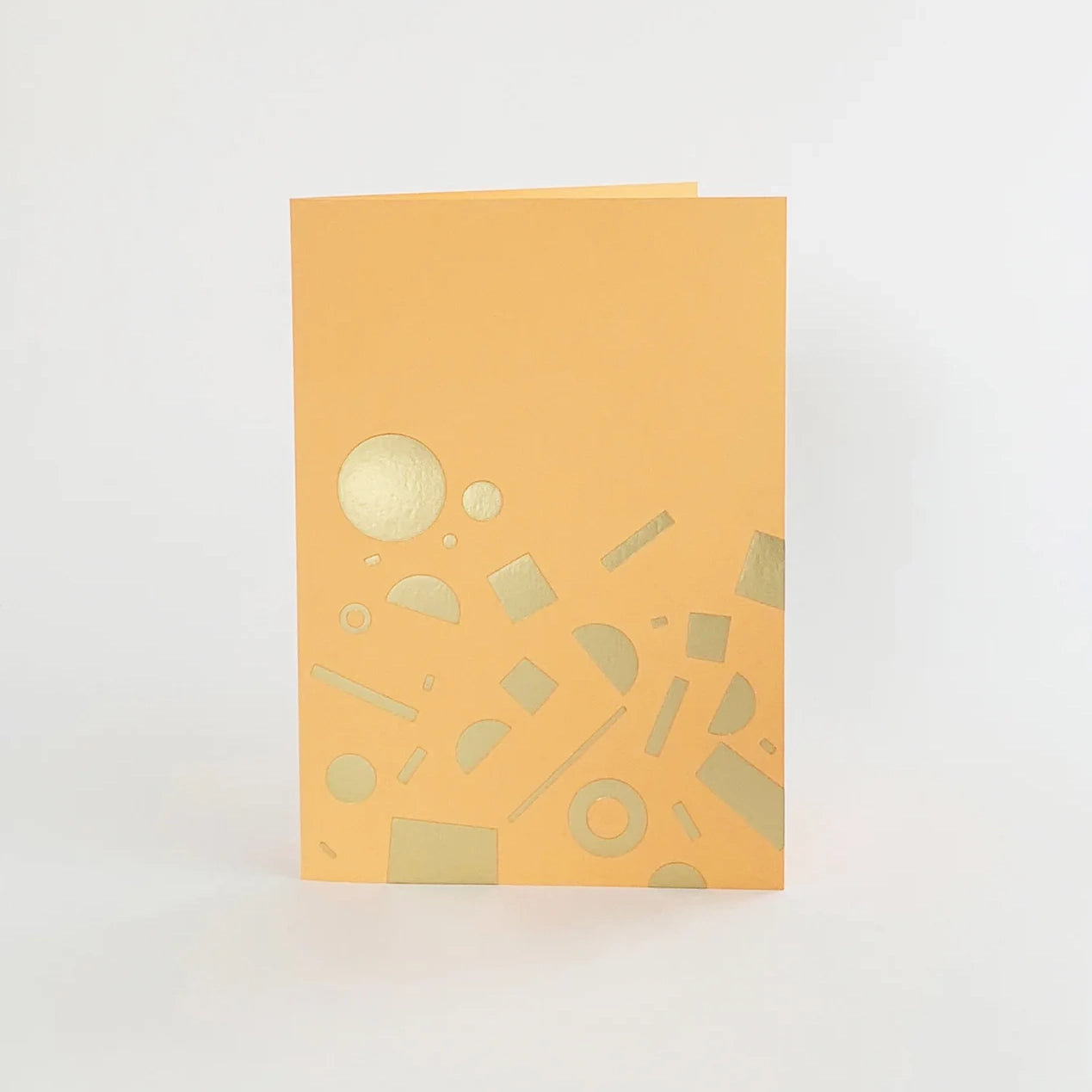 A peach-coloured greeting card with gold foil geometric shapes, including circles, rectangles, and lines, scattered across the card.